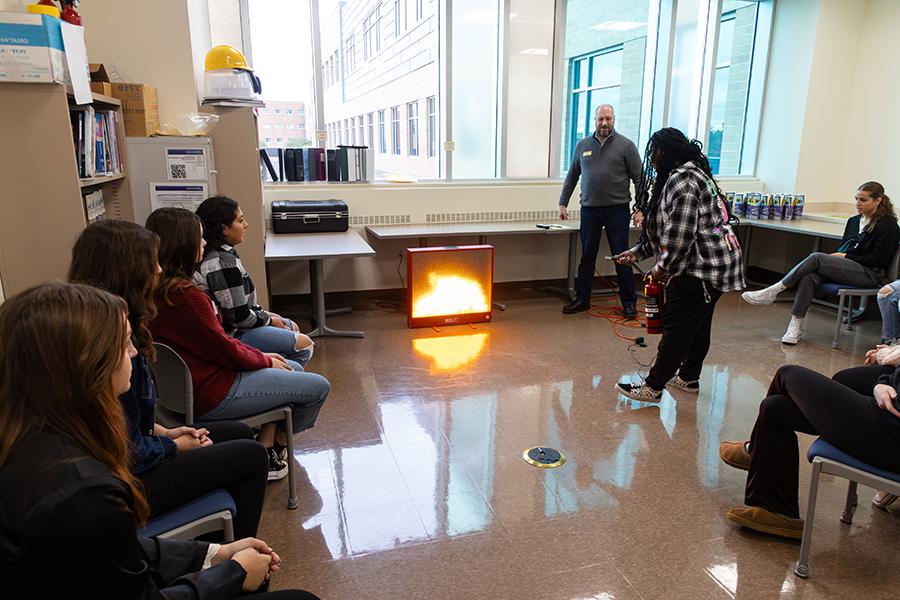 Students are gathered in a classroom with a simulated fireplace and one student is holding a fire extinguisher.